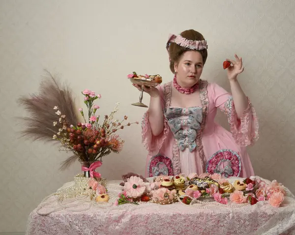 Zengin pembe bir elbise giyen sevimli bir kadın modelin portresi. Tarihi bir Fransız barok asilzadesinin kostümü. Hamur işleri, çiçekler ve zengin mücevherlerle dolu bir masada, hoş bir ziyafette yemek ve tatlı yemek yemek..
