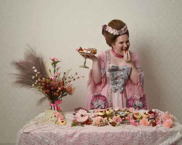 Zengin pembe bir elbise giyen sevimli bir kadın modelin portresi. Tarihi bir Fransız barok asilzadesinin kostümü. Hamur işleri, çiçekler ve zengin mücevherlerle dolu bir masada, hoş bir ziyafette yemek ve tatlı yemek yemek..