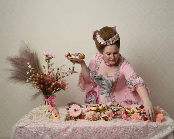 Zengin pembe bir elbise giyen sevimli bir kadın modelin portresi. Tarihi bir Fransız barok asilzadesinin kostümü. Hamur işleri, çiçekler ve zengin mücevherlerle dolu bir masada, hoş bir ziyafette yemek ve tatlı yemek yemek..