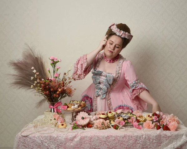Zengin pembe bir elbise giyen sevimli bir kadın modelin portresi. Tarihi bir Fransız barok asilzadesinin kostümü. Hamur işleri, çiçekler ve zengin mücevherlerle dolu bir masada, hoş bir ziyafette yemek ve tatlı yemek yemek..