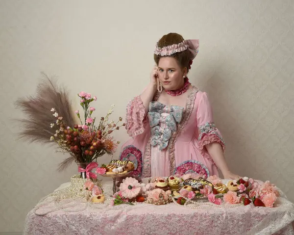 Zengin pembe bir elbise giyen sevimli bir kadın modelin portresi. Tarihi bir Fransız barok asilzadesinin kostümü. Hamur işleri, çiçekler ve zengin mücevherlerle dolu bir masada, hoş bir ziyafette yemek ve tatlı yemek yemek..