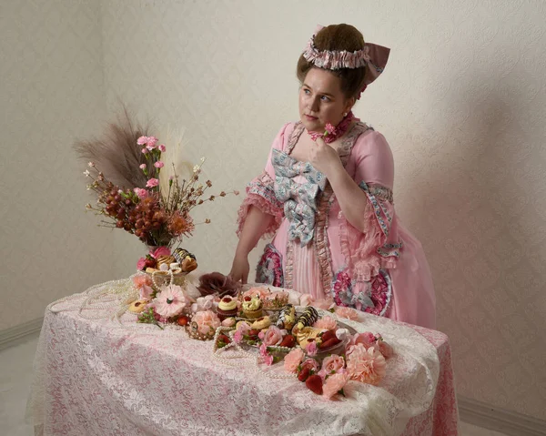 Zengin pembe bir elbise giyen sevimli bir kadın modelin portresi. Tarihi bir Fransız barok asilzadesinin kostümü. Hamur işleri, çiçekler ve zengin mücevherlerle dolu bir masada, hoş bir ziyafette yemek ve tatlı yemek yemek..