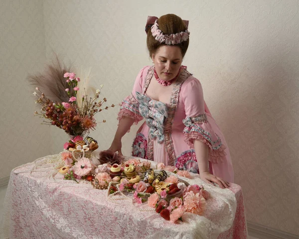 Zengin pembe bir elbise giyen sevimli bir kadın modelin portresi. Tarihi bir Fransız barok asilzadesinin kostümü. Hamur işleri, çiçekler ve zengin mücevherlerle dolu bir masada, hoş bir ziyafette yemek ve tatlı yemek yemek..