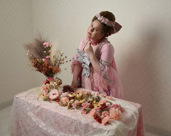 Zengin pembe bir elbise giyen sevimli bir kadın modelin portresi. Tarihi bir Fransız barok asilzadesinin kostümü. Hamur işleri, çiçekler ve zengin mücevherlerle dolu bir masada, hoş bir ziyafette yemek ve tatlı yemek yemek..