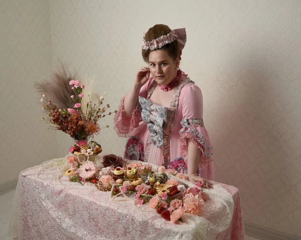 Zengin pembe bir elbise giyen sevimli bir kadın modelin portresi. Tarihi bir Fransız barok asilzadesinin kostümü. Hamur işleri, çiçekler ve zengin mücevherlerle dolu bir masada, hoş bir ziyafette yemek ve tatlı yemek yemek..