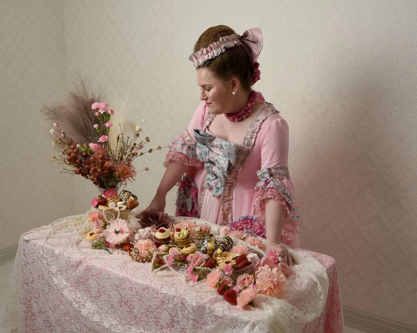 Zengin pembe bir elbise giyen sevimli bir kadın modelin portresi. Tarihi bir Fransız barok asilzadesinin kostümü. Hamur işleri, çiçekler ve zengin mücevherlerle dolu bir masada, hoş bir ziyafette yemek ve tatlı yemek yemek..