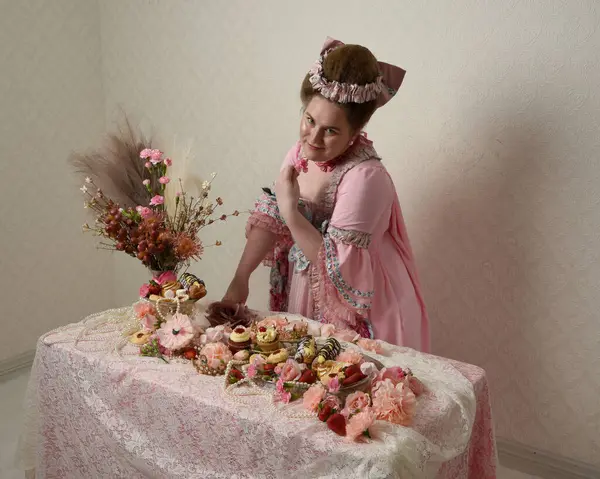 Zengin pembe bir elbise giyen sevimli bir kadın modelin portresi. Tarihi bir Fransız barok asilzadesinin kostümü. Hamur işleri, çiçekler ve zengin mücevherlerle dolu bir masada, hoş bir ziyafette yemek ve tatlı yemek yemek..