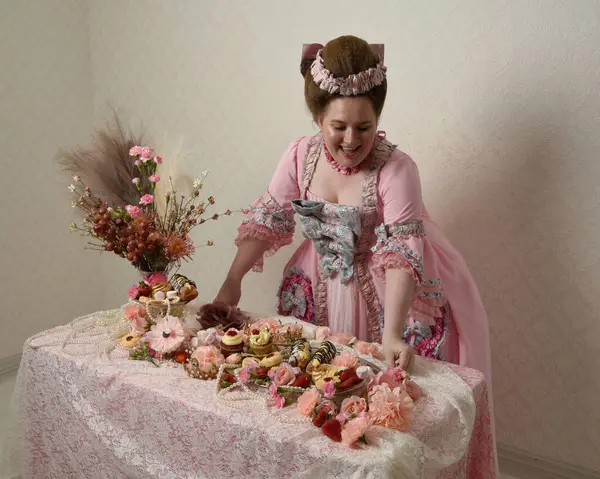 Zengin pembe bir elbise giyen sevimli bir kadın modelin portresi. Tarihi bir Fransız barok asilzadesinin kostümü. Hamur işleri, çiçekler ve zengin mücevherlerle dolu bir masada, hoş bir ziyafette yemek ve tatlı yemek yemek..