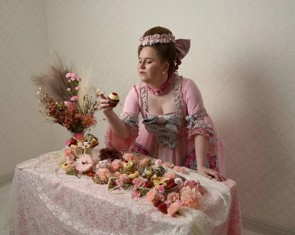 Zengin pembe bir elbise giyen sevimli bir kadın modelin portresi. Tarihi bir Fransız barok asilzadesinin kostümü. Hamur işleri, çiçekler ve zengin mücevherlerle dolu bir masada, hoş bir ziyafette yemek ve tatlı yemek yemek..