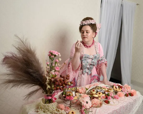 Zengin pembe bir elbise giyen sevimli bir kadın modelin portresi. Tarihi bir Fransız barok asilzadesinin kostümü. Hamur işleri, çiçekler ve zengin mücevherlerle dolu bir masada, hoş bir ziyafette yemek ve tatlı yemek yemek..