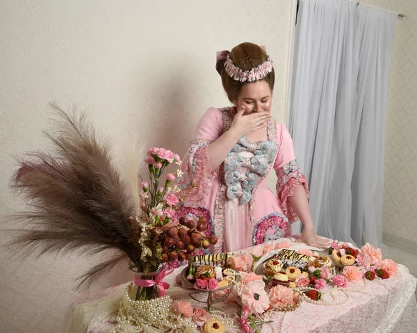 Zengin pembe bir elbise giyen sevimli bir kadın modelin portresi. Tarihi bir Fransız barok asilzadesinin kostümü. Hamur işleri, çiçekler ve zengin mücevherlerle dolu bir masada, hoş bir ziyafette yemek ve tatlı yemek yemek..