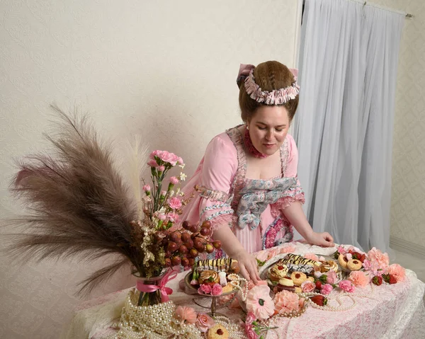 Zengin pembe bir elbise giyen sevimli bir kadın modelin portresi. Tarihi bir Fransız barok asilzadesinin kostümü. Hamur işleri, çiçekler ve zengin mücevherlerle dolu bir masada, hoş bir ziyafette yemek ve tatlı yemek yemek..