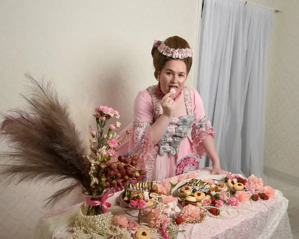 Zengin pembe bir elbise giyen sevimli bir kadın modelin portresi. Tarihi bir Fransız barok asilzadesinin kostümü. Hamur işleri, çiçekler ve zengin mücevherlerle dolu bir masada, hoş bir ziyafette yemek ve tatlı yemek yemek..