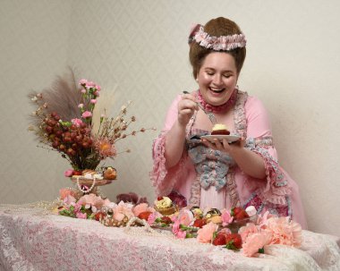 Zengin pembe bir elbise giyen sevimli bir kadın modelin portresi. Tarihi bir Fransız barok asilzadesinin kostümü. Hoşgörülü bir ziyafette tatlı ve zengin yiyeceklerle pasta yemek..