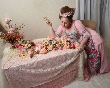 Zengin pembe bir elbise giyen sevimli bir kadın modelin portresi. Tarihi bir Fransız barok asilzadesinin kostümü. Hoş bir ziyafette pasta yemek tatlı ikramları ve arka planda karmaşık duvar kağıtları olan uzun masada zengin yiyecekler yemek..