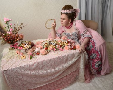 Zengin pembe bir elbise giyen sevimli bir kadın modelin portresi. Tarihi bir Fransız barok asilzadesinin kostümü. Hoş bir ziyafette pasta yemek tatlı ikramları ve arka planda karmaşık duvar kağıtları olan uzun masada zengin yiyecekler yemek..
