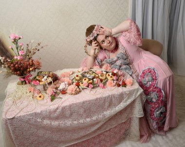 Zengin pembe bir elbise giyen sevimli bir kadın modelin portresi. Tarihi bir Fransız barok asilzadesinin kostümü. Hoş bir ziyafette pasta yemek tatlı ikramları ve arka planda karmaşık duvar kağıtları olan uzun masada zengin yiyecekler yemek..