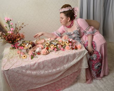 Zengin pembe bir elbise giyen sevimli bir kadın modelin portresi. Tarihi bir Fransız barok asilzadesinin kostümü. Hoş bir ziyafette pasta yemek tatlı ikramları ve arka planda karmaşık duvar kağıtları olan uzun masada zengin yiyecekler yemek..