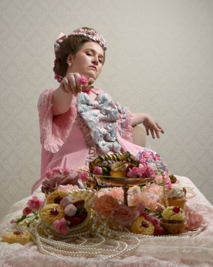 Zengin pembe bir elbise giyen sevimli bir kadın modelin portresi. Tarihi bir Fransız barok asilzadesinin kostümü. Tatlı kekler ve hoşgörülü yemek ziyafetiyle masada oturuyor..