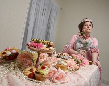 Zengin pembe bir elbise giyen sevimli bir kadın modelin portresi. Tarihi bir Fransız barok asilzadesinin kostümü. Tatlı kekler ve hoşgörülü yemek ziyafetiyle masada oturuyor..