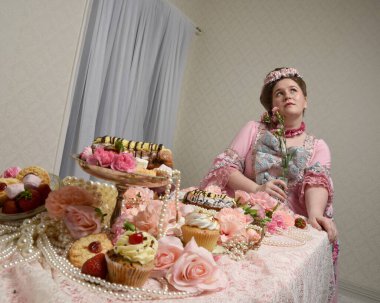 Zengin pembe bir elbise giyen sevimli bir kadın modelin portresi. Tarihi bir Fransız barok asilzadesinin kostümü. Tatlı kekler ve hoşgörülü yemek ziyafetiyle masada oturuyor..