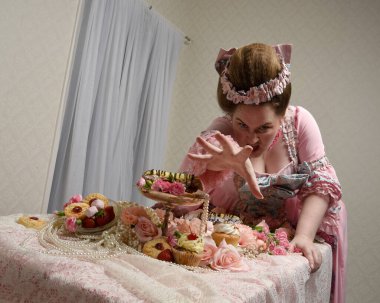 Zengin pembe bir elbise giyen sevimli bir kadın modelin portresi. Tarihi bir Fransız barok asilzadesinin kostümü. Tatlı kekler ve hoşgörülü yemek ziyafetiyle masada oturuyor..