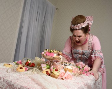 Zengin pembe bir elbise giyen sevimli bir kadın modelin portresi. Tarihi bir Fransız barok asilzadesinin kostümü. Tatlı kekler ve hoşgörülü yemek ziyafetiyle masada oturuyor..