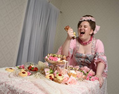 Zengin pembe bir elbise giyen sevimli bir kadın modelin portresi. Tarihi bir Fransız barok asilzadesinin kostümü. Tatlı kekler ve hoşgörülü yemek ziyafetiyle masada oturuyor..