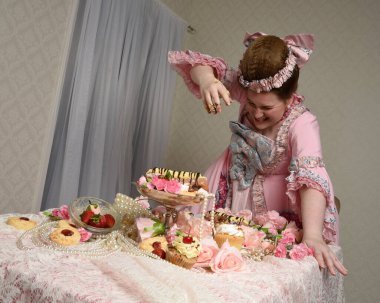 Zengin pembe bir elbise giyen sevimli bir kadın modelin portresi. Tarihi bir Fransız barok asilzadesinin kostümü. Tatlı kekler ve hoşgörülü yemek ziyafetiyle masada oturuyor..