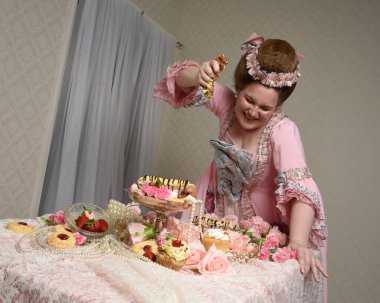Zengin pembe bir elbise giyen sevimli bir kadın modelin portresi. Tarihi bir Fransız barok asilzadesinin kostümü. Tatlı kekler ve hoşgörülü yemek ziyafetiyle masada oturuyor..