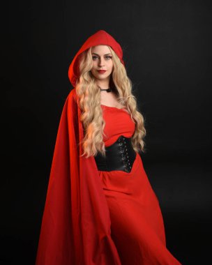 Güzel bir mankenin portresi, uzun sarı saçlar, göz alıcı fantezi elbiseler, korse ve akışkan kırmızı başlıklı peri masalı pelerini. Karanlık stüdyo arka planında izole.