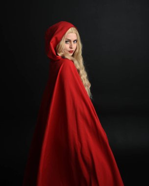 Güzel bir mankenin portresi, uzun sarı saçlar, göz alıcı fantezi elbiseler, korse ve akışkan kırmızı başlıklı peri masalı pelerini. Karanlık stüdyo arka planında izole.