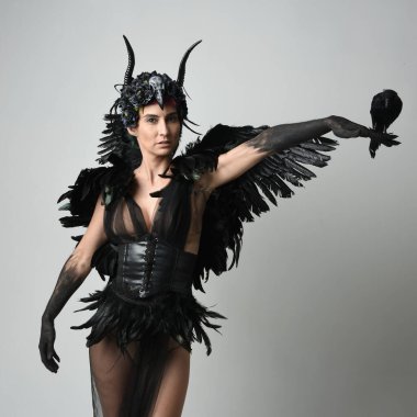 Gotik boynuzlu başlıklı, cadılar bayramı siyah elbiseli ve fantezi melek kanatlı kadın modelin yakın portresi. El kol pozu veren bir kuş figürü. İzole stüdyo geçmişi