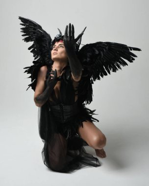 Gotik boynuzlu başlıklı, cadılar bayramı siyah elbiseli ve fantezi melek kanatlı kadın modelin tam boy portresi. Çömelme pozisyonu, diz çökme. İzole stüdyo geçmişi