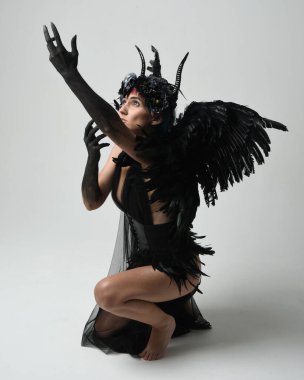 Gotik boynuzlu başlıklı, cadılar bayramı siyah elbiseli ve fantezi melek kanatlı kadın modelin tam boy portresi. Çömelme pozisyonu, diz çökme. İzole stüdyo geçmişi