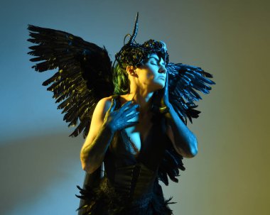 Gotik boynuzlu başlık giyen kadın modelin yakın portresi, cadılar bayramı siyah elbisesi ve fantezi melek kanatları. Sinema gölge siluetleriyle izole edilmiş karanlık stüdyo arkaplanı 