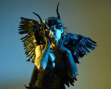 Gotik boynuzlu başlık giyen kadın modelin yakın portresi, cadılar bayramı siyah elbisesi ve fantezi melek kanatları. Sinema gölge siluetleriyle izole edilmiş karanlık stüdyo arkaplanı 