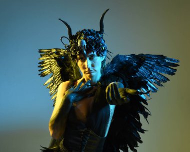 Gotik boynuzlu başlık giyen kadın modelin yakın portresi, cadılar bayramı siyah elbisesi ve fantezi melek kanatları. Sinema gölge siluetleriyle izole edilmiş karanlık stüdyo arkaplanı 