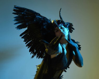 Gotik boynuzlu başlık giyen kadın modelin yakın portresi, cadılar bayramı siyah elbisesi ve fantezi melek kanatları. Sinema gölge siluetleriyle izole edilmiş karanlık stüdyo arkaplanı 