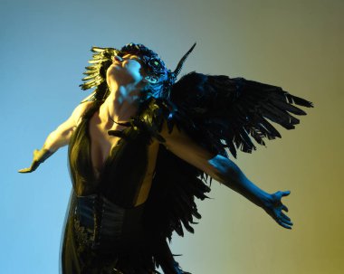 Gotik boynuzlu başlık giyen kadın modelin yakın portresi, cadılar bayramı siyah elbisesi ve fantezi melek kanatları. Sinema gölge siluetleriyle izole edilmiş karanlık stüdyo arkaplanı 