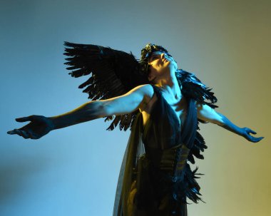 Gotik boynuzlu başlık giyen kadın modelin yakın portresi, cadılar bayramı siyah elbisesi ve fantezi melek kanatları. Sinema gölge siluetleriyle izole edilmiş karanlık stüdyo arkaplanı 