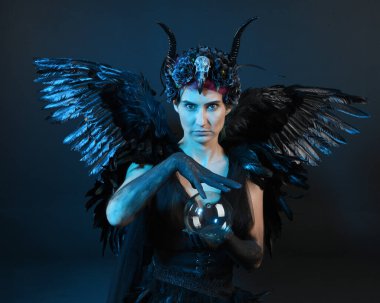 Gotik boynuzlu başlık giyen kadın modelin yakın portresi, cadılar bayramı siyah elbisesi, fantezi melek tüyü kanatları. Mistik kristal küreyi tutuyorum. Karanlık stüdyo arka planı, sinematik gölge silueti 