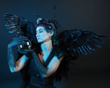 Gotik boynuzlu başlık giyen kadın modelin yakın portresi, cadılar bayramı siyah elbisesi, fantezi melek tüyü kanatları. Mistik kristal küreyi tutuyorum. Karanlık stüdyo arka planı, sinematik gölge silueti 