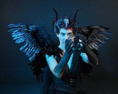 Gotik boynuzlu başlık giyen kadın modelin yakın portresi, cadılar bayramı siyah elbisesi, fantezi melek tüyü kanatları. Mistik kristal küreyi tutuyorum. Karanlık stüdyo arka planı, sinematik gölge silueti 