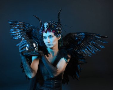 Gotik boynuzlu başlık giyen kadın modelin yakın portresi, cadılar bayramı siyah elbisesi, fantezi melek tüyü kanatları. Mistik kristal küreyi tutuyorum. Karanlık stüdyo arka planı, sinematik gölge silueti 