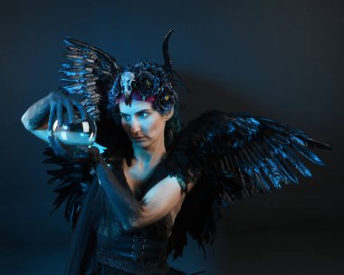 Gotik boynuzlu başlık giyen kadın modelin yakın portresi, cadılar bayramı siyah elbisesi, fantezi melek tüyü kanatları. Mistik kristal küreyi tutuyorum. Karanlık stüdyo arka planı, sinematik gölge silueti 