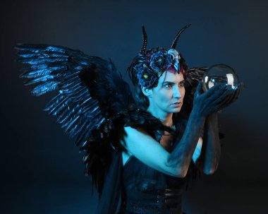 Gotik boynuzlu başlık giyen kadın modelin yakın portresi, cadılar bayramı siyah elbisesi, fantezi melek tüyü kanatları. Mistik kristal küreyi tutuyorum. Karanlık stüdyo arka planı, sinematik gölge silueti 