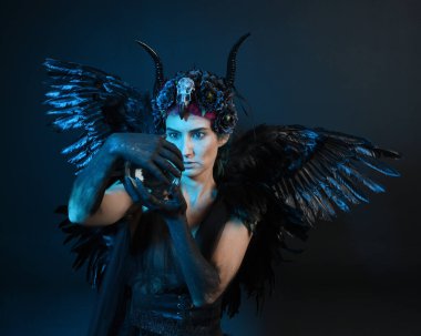 Gotik boynuzlu başlık giyen kadın modelin yakın portresi, cadılar bayramı siyah elbisesi, fantezi melek tüyü kanatları. Mistik kristal küreyi tutuyorum. Karanlık stüdyo arka planı, sinematik gölge silueti 