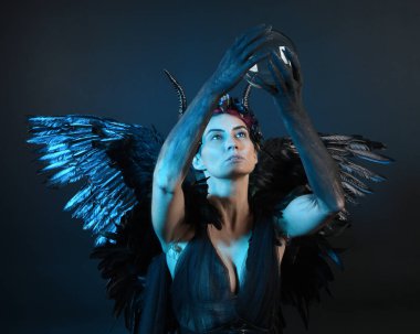 Gotik boynuzlu başlık giyen kadın modelin yakın portresi, cadılar bayramı siyah elbisesi, fantezi melek tüyü kanatları. Mistik kristal küreyi tutuyorum. Karanlık stüdyo arka planı, sinematik gölge silueti 