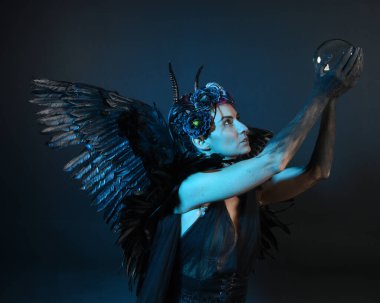 Gotik boynuzlu başlık giyen kadın modelin yakın portresi, cadılar bayramı siyah elbisesi, fantezi melek tüyü kanatları. Mistik kristal küreyi tutuyorum. Karanlık stüdyo arka planı, sinematik gölge silueti 